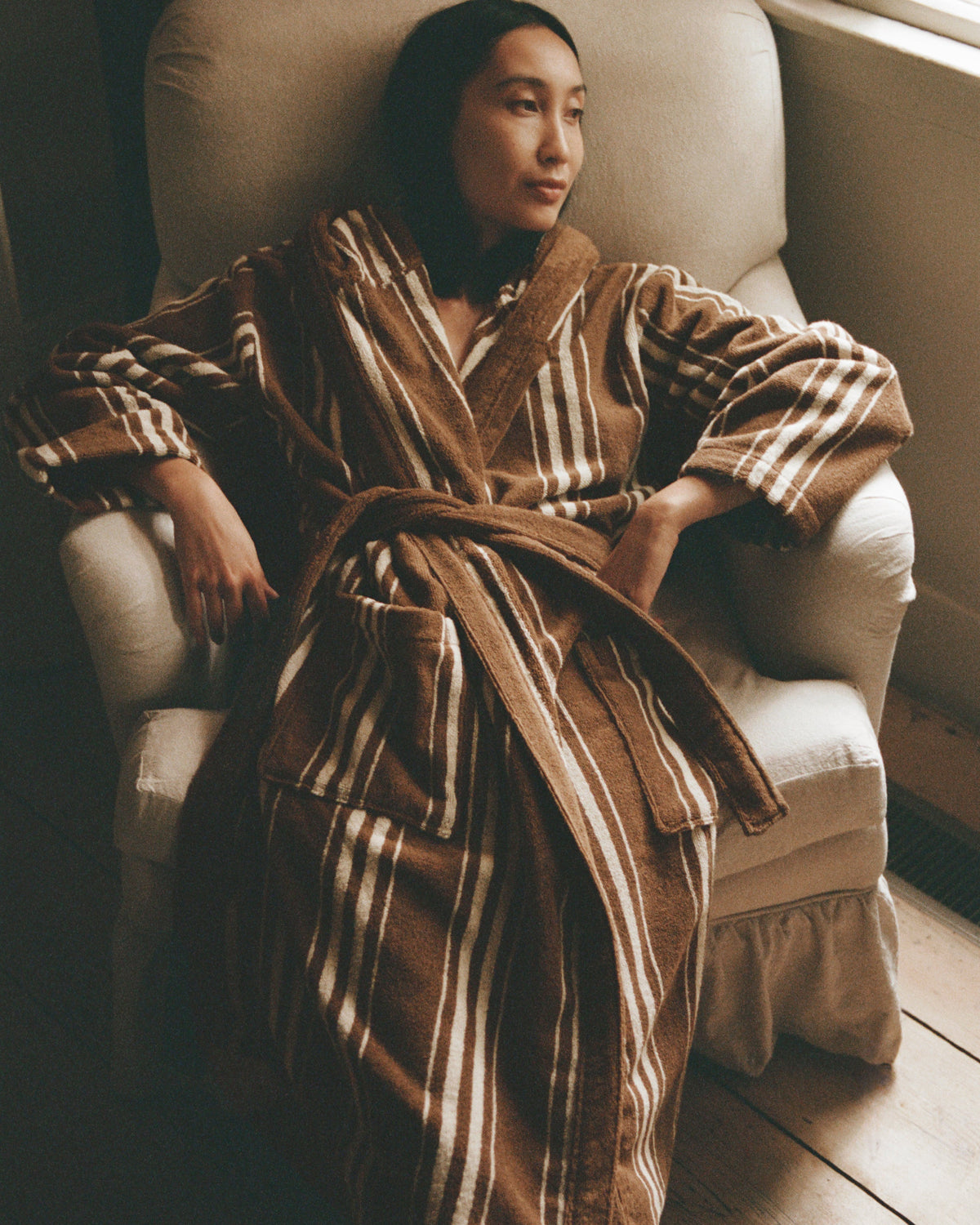 A woman wearing a brown and beige striped plush bathrobe sits comfortably in a light-colored armchair on a wooden floor.