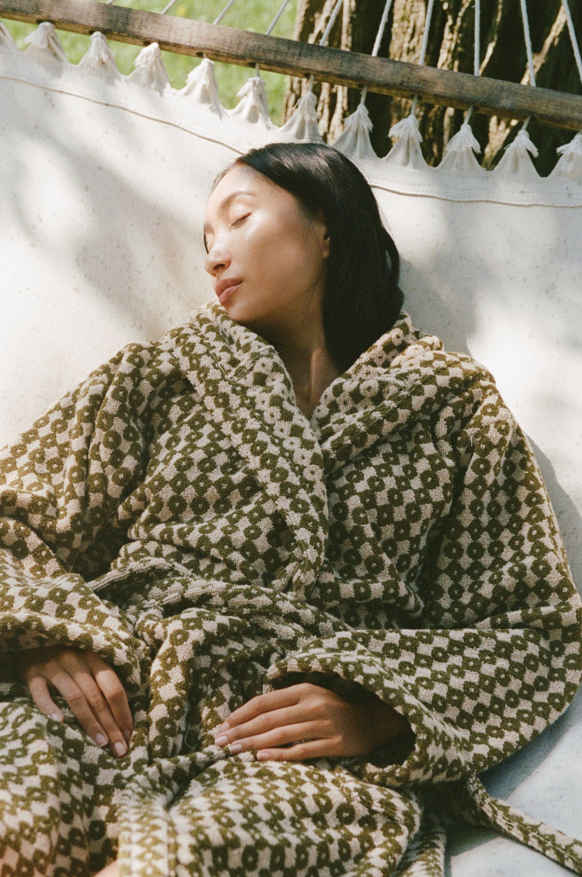 A woman reclining outdoors on a white hammock, wrapped in a plush robe with a geometric pattern.